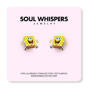 May include: A pair of yellow SpongeBob SquarePants earrings with brown pants and black shoes. The earrings are on a pink background with the text "SOUL WHISPERS JEWELRY". The earrings are made with hypo-allergenic stainless steel posts and backs.
