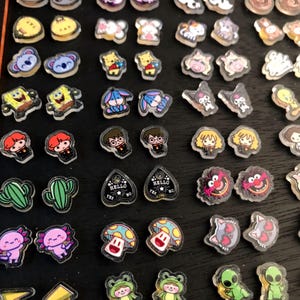 May include: A collection of clear, resin-like charms with various cartoon and character designs. The charms feature images of koalas, Winnie the Pooh, SpongeBob, Harry Potter, and more. Some charms have the text "HELLO YES NO".