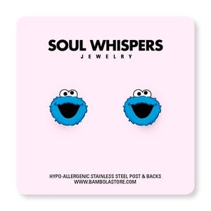 May include: A pair of blue Cookie Monster stud earrings with black mouths and white eyes. The earrings are displayed on a pink background with the text "SOUL WHISPERS JEWELRY". The text "HYPO-ALLERGENIC STAINLESS STEEL POST & BACKS" is also visible.