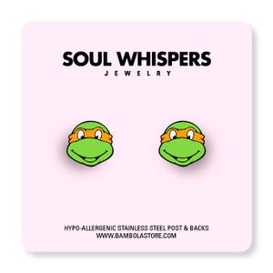 May include: A pair of stud earrings featuring the face of a Teenage Mutant Ninja Turtle with a green face and orange mask. The earrings are on a pink card with the text "SOUL WHISPERS JEWELRY" and "HYPO-ALLERGENIC STAINLESS STEEL POST & BACKS".