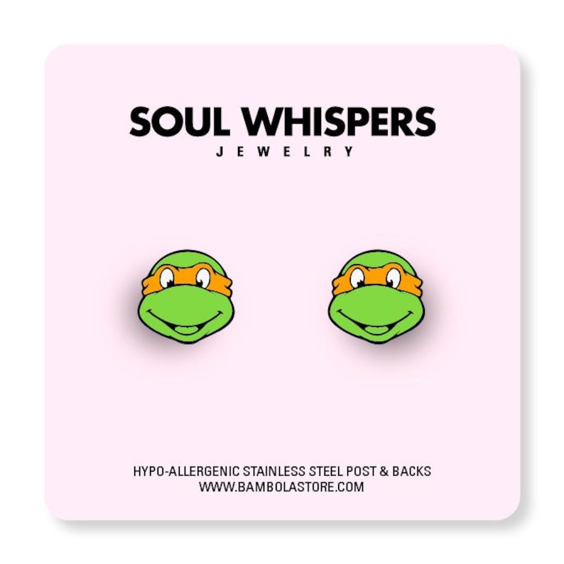 May include: A pair of stud earrings featuring the face of a Teenage Mutant Ninja Turtle with a green face and orange mask. The earrings are on a pink card with the text "SOUL WHISPERS JEWELRY" and "HYPO-ALLERGENIC STAINLESS STEEL POST & BACKS".