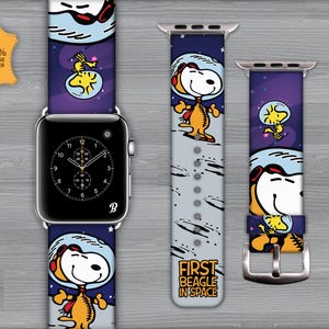 Puede incluir: Una correa de reloj con un diseño espacial que presenta a Snoopy en un traje espacial. La correa es blanca con detalles en negro y gris y el texto "First Beagle in Space". La esfera del reloj es negra con manecillas y números blancos.