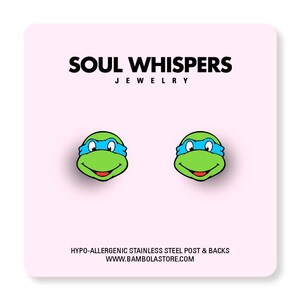 May include: A pair of green and blue cartoon character earrings. The earrings are shaped like a turtle's face with a blue mask. The background is a light pink card with the text "SOUL WHISPERS JEWELRY". The text "HYPO-ALLERGENIC STAINLESS STEEL POST & BACKS" is also visible.