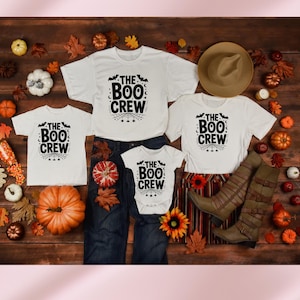 May include: A Halloween-themed flat lay featuring white t-shirts and a onesie, all printed with "The Boo Crew" in black. The shirts are arranged with a pair of blue jeans, a brown hat, and tall brown boots. Pumpkins and autumn leaves complete the festive scene.