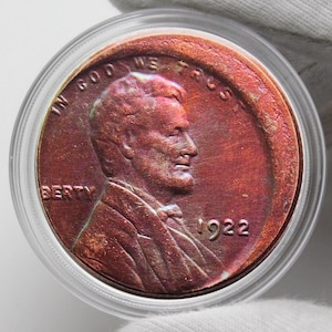 Puede incluir: Una moneda de cobre de 1922 dentro de un soporte de plástico transparente. La moneda presenta el perfil de un hombre y las palabras "IN GOD WE TRUST" y "LIBERTY". El año "1922" es visible.