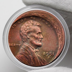 Puede incluir: Un centavo Lincoln de 1951, de color cobre, en una funda protectora transparente. La moneda presenta un perfil de Abraham Lincoln y las inscripciones "IN GOD WE TRUST" y "LIBERTY". El año "1951" también es visible.