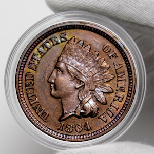 May include: A copper Indian Head penny from 1864, in a clear protective case. The coin shows a profile of a Native American in a feathered headdress and the words "UNITED STATES OF AMERICA".