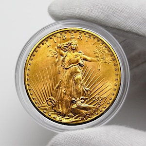 Op de afbeelding: Een gouden munt met een afbeelding van een vrouw die een fakkel en een tak met olijfbladeren vasthoudt. De munt heeft de inscripties "LIBERTY" en "1927".