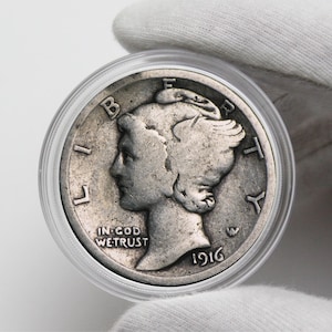 May include: A silver 1916 Walking Liberty Half Dollar coin in a plastic coin holder. The coin features a profile of Liberty walking with a torch and an olive branch. The inscription "IN GOD WE TRUST" is visible below Liberty's image.