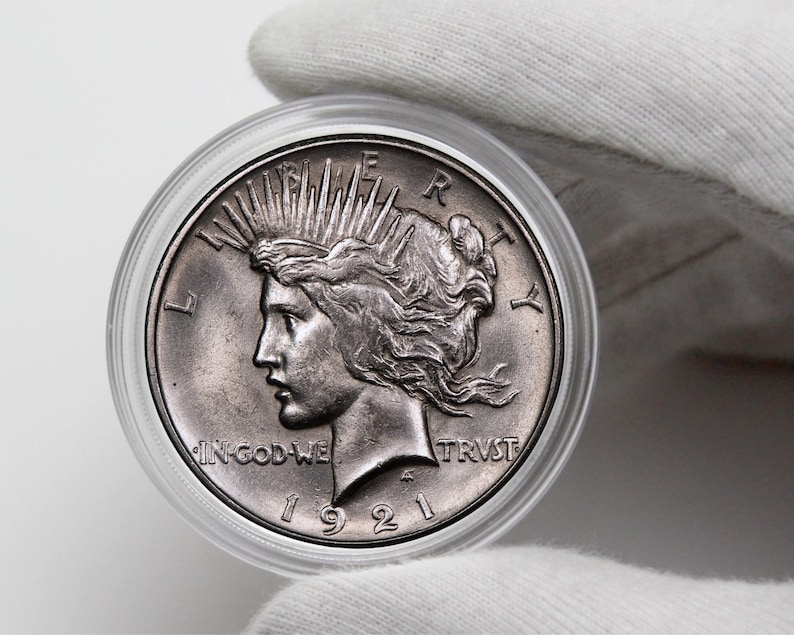 May include: A silver 1921 Peace Dollar coin in a clear plastic coin holder. The coin features a profile of a woman with a crown of rays, the words "LIBERTY" and "IN GOD WE TRUST", and the year "1921".
