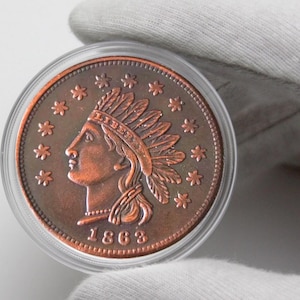 May include: A copper-coloured coin depicting a profile of a Native American in a feathered headdress, encircled by stars. The year 1863 is shown. The coin is in a clear protective case.