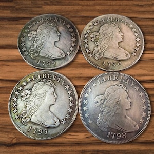 Pode incluir: Quatro moedas antigas de prata com o perfil de uma mulher, a palavra "LIBERTY" e os anos 1795, 1796, 1797 e 1798. As moedas estão dispostas sobre uma superfície de madeira.