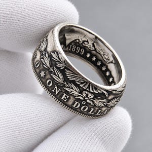Peut inclure: Une bague en forme de pièce de monnaie argentée fabriquée à partir d'un dollar américain vintage, présentant des détails en relief visibles, notamment le texte ONE DOLLAR, une étoile, des branches de laurier et l'année 1899 gravée.