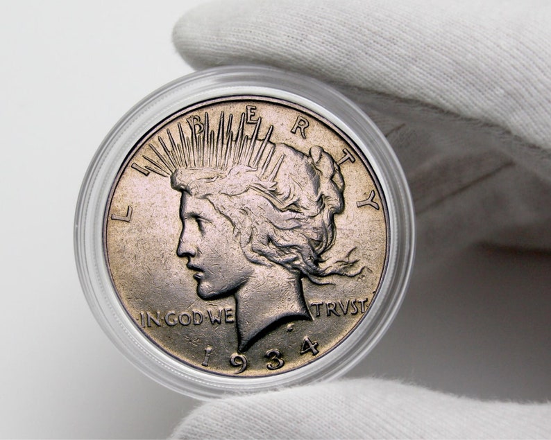 Op de afbeelding: Een zilveren Peace Dollar-munt uit 1934, met een profiel van Liberty met stralen licht achter haar hoofd, de woorden "IN GOD WE TRUST" en "LIBERTY" op de munt, en de datum "1934" eronder.