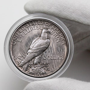 May include: A silver Peace Dollar coin in a clear plastic case. The coin features a bald eagle with outstretched wings, the words "United States of America" and "E Pluribus Unum", and the inscription "One Dollar" and "Peace".