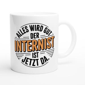 Könnte beinhalten: Weiße Keramik-Tasse mit gebogenem Henkel. Die Tasse hat ein kreisförmiges Design mit dem Text "ALLES WIRD GUT DER INTERNIST IST JETZT DA" in schwarzer und orangefarbener Schrift.