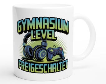 Könnte beinhalten: Weiße Keramik-Tasse mit Gaming-Design. Das Design zeigt Kopfhörer, einen Gamecontroller und den Text "GYMNASIUM LEVEL FREIGESCHALTET" in Blau und Grün. Die Tasse hat einen gebogenen Griff.