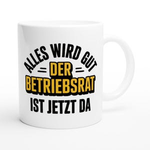 Könnte beinhalten: Weißer Keramikbecher mit gebogenem Henkel. Der Becher hat einen schwarzen und gelben Text mit der Aufschrift "ALLES WIRD GUT = DER BETRIEBSRAT IST JETZT DA". Der Text ist in einem Halbkreis angeordnet.