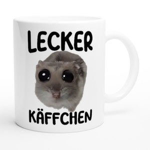 Könnte beinhalten: Weiße Keramiktasse mit einem Schwarzweißbild eines Hamsters mit großen Augen. Der Text "Lecker Käffchen" ist auf die Tasse gedruckt.