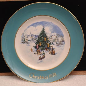 May include: A round ceramic collector plate with a teal border and gold rim. The center features a snowy scene of people decorating a Christmas tree in a village. The text "Christmas 1978" is printed in gold at the bottom of the teal border.