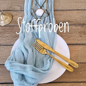 Könnte beinhalten: Ein hellblauer Stoffläufer mit einem goldenen geometrischen Kerzenhalter und einer weißen Kerze. Der Läufer liegt auf einem Holztisch mit einem weißen Teller und goldenem Besteck. Der Text "Stoffproben" ist auf dem Läufer geschrieben.