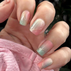May include: A hand displaying a set of square-shaped artificial nails. The nails feature a gradient design blending pink, green, and white hues, accented with a delicate gold glitter line. The nails are set against a soft pink fabric background.
