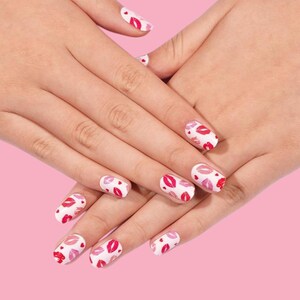 May include: A pair of hands with manicured nails. Each nail is painted white and decorated with a pattern of red and pink lips. The background is a solid pink color.