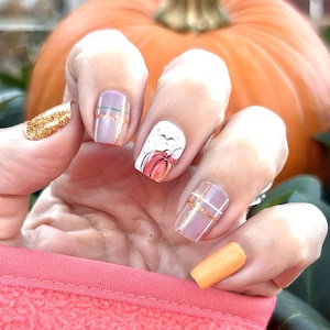 May include: A hand displays a set of fall-themed nail art. The nails feature designs including gold glitter, a pumpkin illustration, and gold accent lines on a light purple base. An orange pumpkin is in the background.