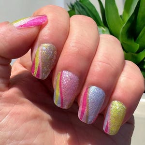 May include: Close-up of a hand with manicured nails. The nails are painted with a glittery finish in shades of pink, yellow, and silver. Each nail features a vertical stripe design.