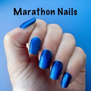 May include: Hand with blue nail polish. The nails are a vibrant, glossy blue color. The text "Marathon Nails" is displayed in black, cursive font at the top of the image.