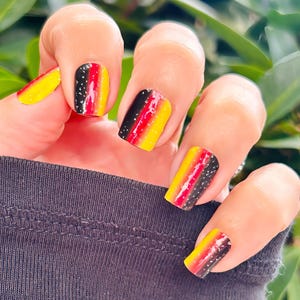 May include: Close-up of a hand with black, yellow, and red nail polish. The nails are decorated with a black and white glitter design.