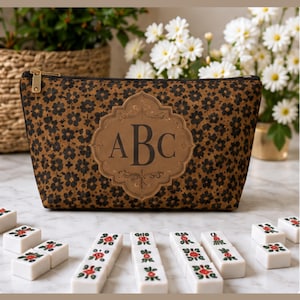 May include: A brown and black floral patterned zippered pouch featuring a central tan decorative emblem with the letters ABC printed in dark brown. The pouch has a gold-toned metal zipper pull and is constructed from a textured material.