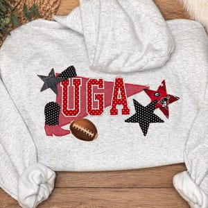 May include: A light grey hoodie with the word "UGA" in red with white polka dots. The design includes a football, stars, and a boot. The hoodie is made of a soft material.