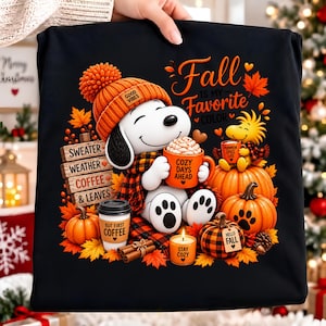 Puede incluir: Una camiseta negra con un gráfico de un perro blanco con gorro de punto naranja y bufanda de cuadros, sosteniendo una taza naranja con 'COZY DAYS AHEAD'. El texto incluye 'Fall IS MY FAVORITE COLOR', 'SWEATER WEATHER COFFEE & LEAVES' y 'BUT FIRST COFFEE'.