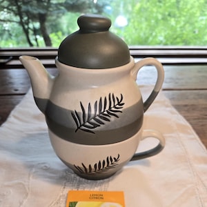 May include: A ceramic tea set with a teapot and teacup, featuring a grey and white striped design and fern illustrations. The set includes a matching lid and is accompanied by three packets of lemon tea.