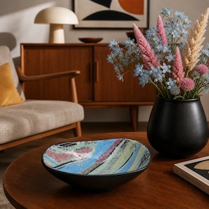 May include: A decorative black ceramic bowl with a colourful, abstract design. The bowl's interior has blue, green, pink, and black brushstrokes. A black vase with flowers and a book titled "German Ceramics" are on a wooden table.