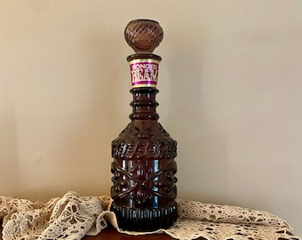 May include: A dark purple glass decanter with a decorative stopper and a label that reads "BONDED BEAM". The decanter is sitting on a wooden shelf with a crocheted lace doily.
