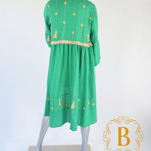 May include: A green dress with gold embroidery and tassels. The dress has a round neckline and a loose fit. It is made of a lightweight fabric and has a flowing silhouette.