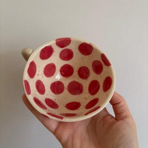 Può includere: Ciotola in ceramica fatta a mano con base color crema e grandi pois rossi. La ciotola, con un piccolo manico, è tenuta in una mano. L'interno della ciotola è anch'esso decorato con pois rossi.