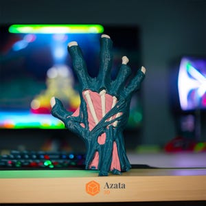 May include: A 3D printed hand model with a teal blue skin and exposed pink bones. The hand is posed with fingers spread apart and is resting on a wooden surface. The text "Azata 3D" is visible in the lower right corner of the image.