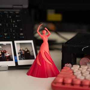 May include: A 3D printed figurine of a person in a red dress with arms raised above their head. The figurine is on a white surface with a laptop and a keyboard in the background.