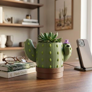 Pode incluir: Um vaso verde em forma de cacto com uma planta suculenta. O vaso tem uma base marrom e é decorado com pequenos detalhes verde claro. Um par de óculos e livros estão na superfície de madeira.