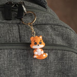 May include: A small orange fox keychain with white accents and big eyes. The keychain is attached to a gold chain and a gold ring.