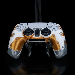 May include: A white and gold custom-designed PlayStation 5 controller with a textured finish. The controller is on a black stand.