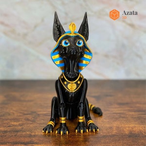 Op de afbeelding: Zwarte kattenfiguur met gouden accenten, die een blauwe en gouden farao-hoofdtooi draagt. Het beeldje heeft blauwe ogen en zit met zijn poten naar voren.
