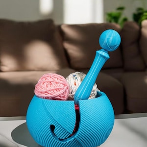 May include: A blue yarn bowl with a yarn holder and a blue yarn needle. The bowl has a textured design and is filled with two balls of yarn, one pink and one white.
