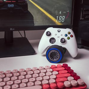 Könnte beinhalten: Ein weißer Xbox-Controller mit einem schwarzen und blauen Lenkrad-Anbauteil. Der Controller liegt auf einer weißen Oberfläche neben einer rot-pinken Tastatur mit runden Tasten. Die Tastatur trägt den Schriftzug "logi". Im Hintergrund ist ein Computermonitor mit einem Rennspiel auf dem Bildschirm zu sehen.