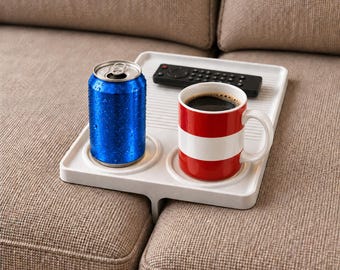 May include: A white plastic couch arm tray with two circular cup holders and a flat surface. A blue aluminum soda can sits in one holder, a red and white striped ceramic mug filled with dark liquid in the other, and a black remote control.