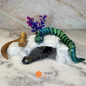 Op de afbeelding: Drie beweeglijke speelgoedslangen in bruin-, groen- en zwarttinten worden getoond op een witte rotsformatie met paars blad. De slangen hebben gedetailleerde ogen en gesegmenteerde lichamen. Het Azata 3D-logo is zichtbaar.