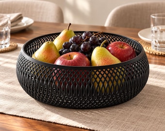 May include: A round black fruit bowl with a decorative lattice pattern containing two yellow and green pears, two red apples, and a cluster of dark purple grapes. The bowl has a wide, open-top design with a structured, repeating geometric weave.